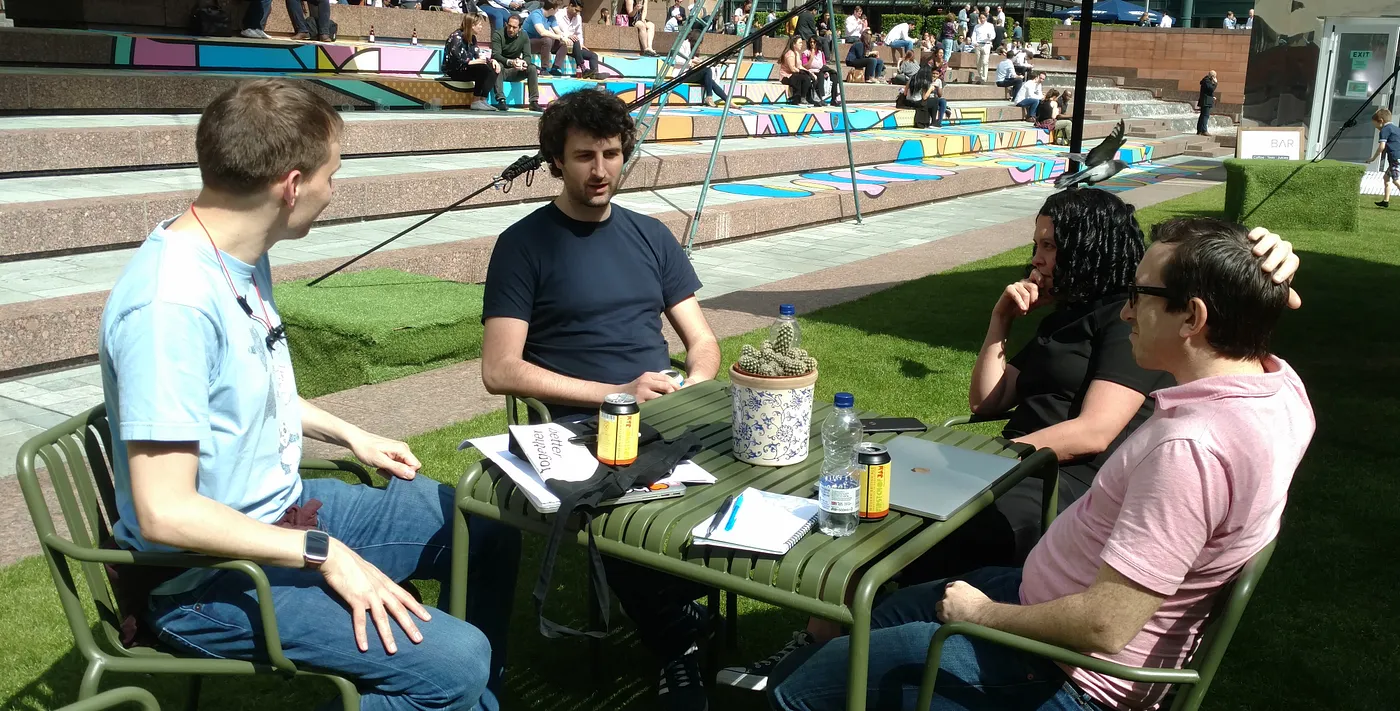 Photo of four Buildsters sitting around a table, outdoors in the sun. One of them is talking and the others are listening intently. There are drinks, laptops and notebooks strewn across the table.